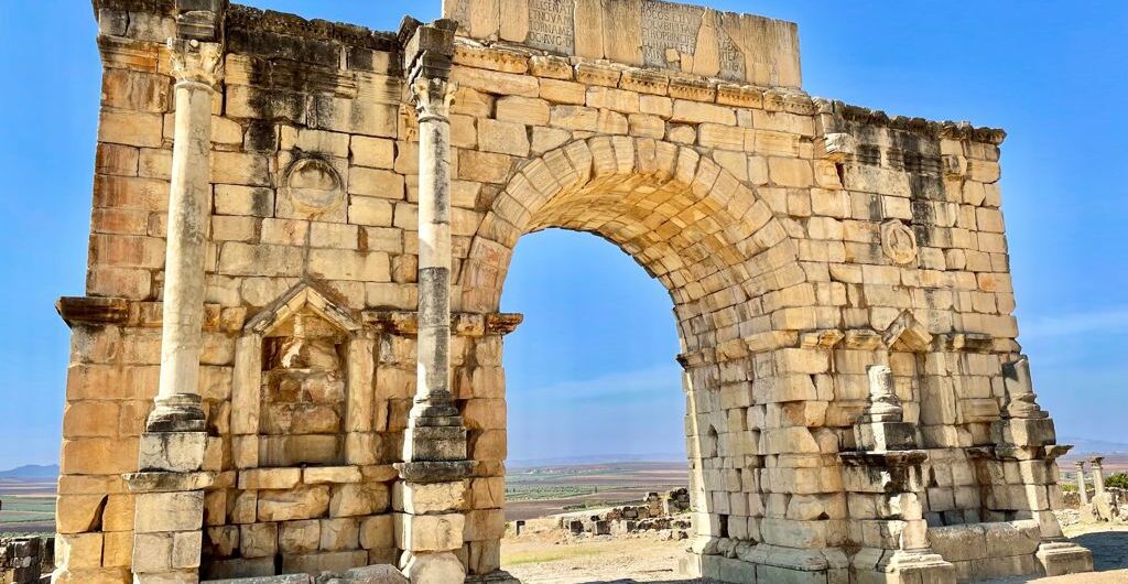 A photograph showcasing the Arch of Triumph in Volubilis, Morocco, standing proudly against a clear blue sky. The ancient stone archway, adorned with intricate carvings, serves as a captivating reminder of Roman architecture and the rich history of the region. #Volubilis #Morocco #Archaeology #Travel